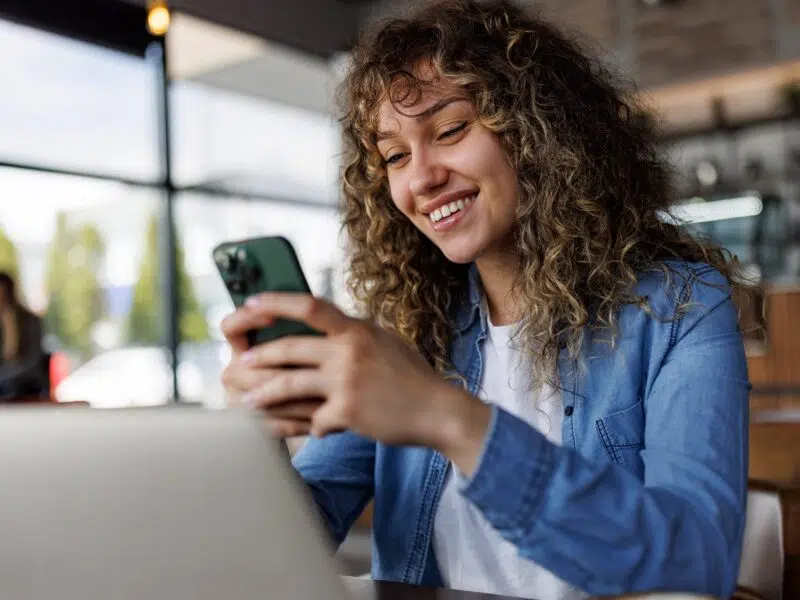 women sitting at laptop inside smiling whilst looking at phone
