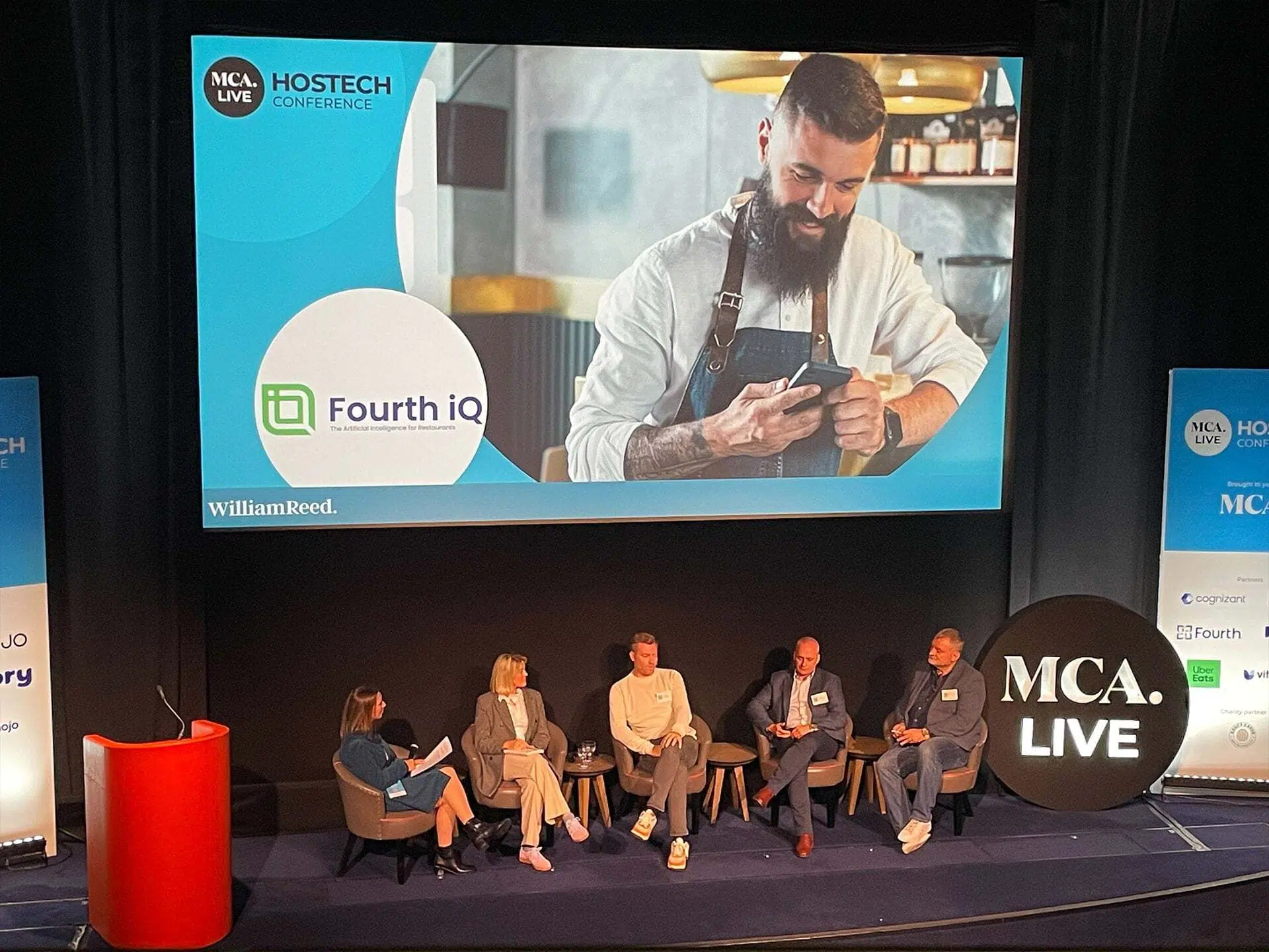 Hostech event with panel speakers on stage and large screen behind with Fourth logo